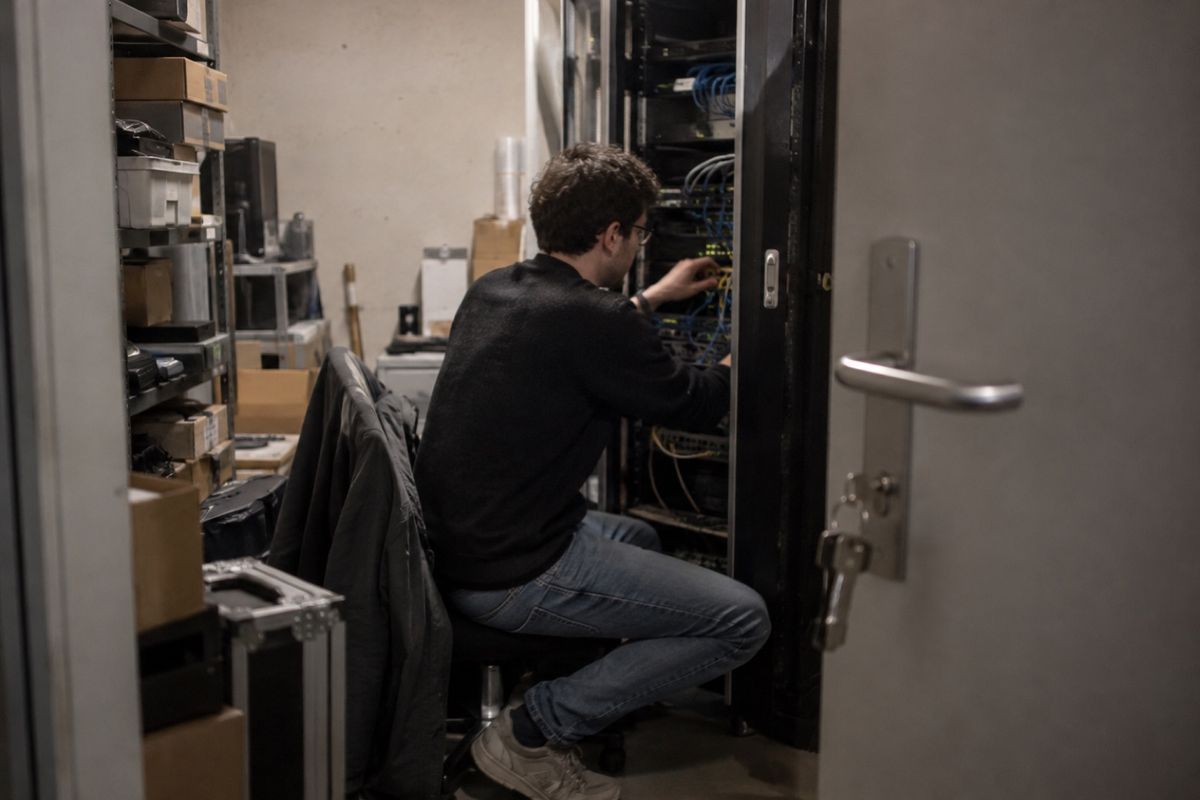 Symbolbild: Ein Mitarbeiter verkabelt in einem engen Technikraum einen offenen Serverschrank, im Vordergrund steht die Tür mit steckendem Schlüssel offen